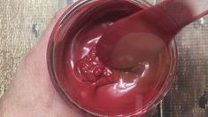 a person holding a spoon in a jar filled with red liquid on top of a wooden table