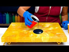 a person in blue gloves pours liquid onto a yellow table top with an orange and black design