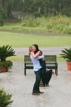 a man and woman hugging each other in front of a bench on a rainy day