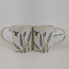 two coffee mugs with blue flowers and a bee painted on the sides, one is white