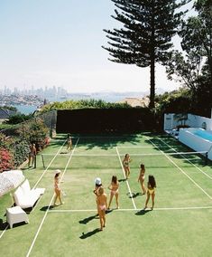 several women in bathing suits playing tennis on a grass court with a view of the city behind them