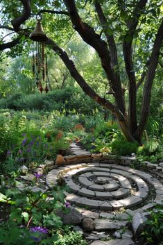 a stone path in the middle of a garden surrounded by trees and flowers with a wind chime hanging from it's center