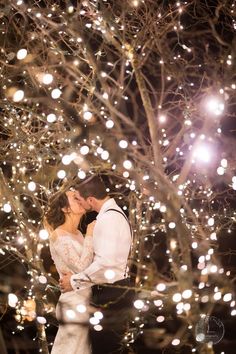 a bride and groom kissing in front of a tree with lights on it at night