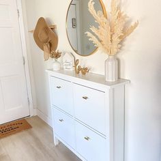 a white dresser topped with a mirror and a vase filled with dried flowers next to a hat on top of it