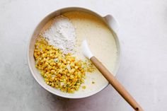 a bowl filled with corn and flour next to a wooden spoon on a white surface