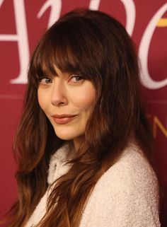 a woman with long hair and bangs standing in front of a red wall wearing a white sweater