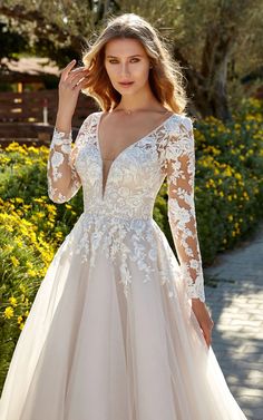 a woman wearing a wedding dress with long sleeves and sheer lace on the top, standing in front of flowers