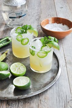 two glasses filled with lemonade and cucumbers on a metal tray next to limes