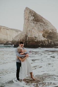 a man and woman hugging in the water at the beach with rocks in the background