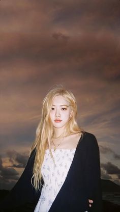 a woman with long blonde hair standing in front of a sky filled with clouds at sunset