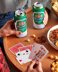 two people playing cards at a table with beer and snacks on it, while another person holds up a deck of cards