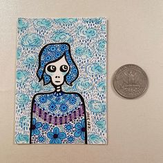 a coin sitting next to a drawing of a woman with blue hair and flowers on it