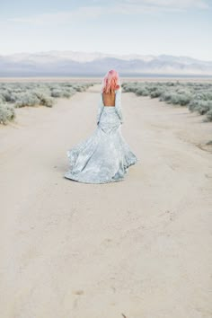 a woman with pink hair is walking in the middle of an empty desert road wearing a silver dress