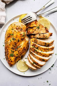 a white plate topped with sliced chicken and lemon wedges next to a knife and fork