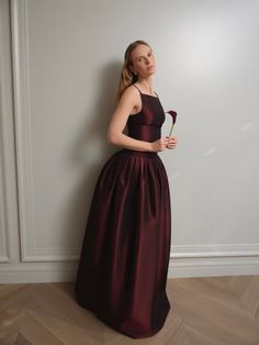 a woman in a long dress leaning against a wall and holding a flower with her right hand