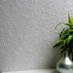 a green plant in a silver vase next to a white wallpapered with flowers