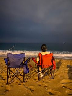 two people sitting in lawn chairs on the beach watching the ocean with dark clouds overhead