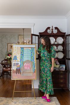 a woman standing in front of a painting next to a wooden cabinet and table with china on it