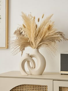 a white vase with dried grass in it on top of a table next to a radio