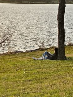 a man sitting under a tree on top of a lush green field next to a lake