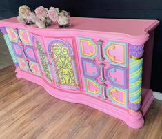 a pink and blue painted dresser with flowers on the top, in front of a black wall