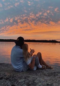 a man and woman are sitting on the shore watching the sunset