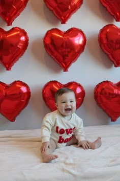 a baby sitting on a bed in front of many red heart shaped balloon wall decorations