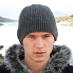 a young man wearing a knitted hat and jacket next to the ocean with a cell phone in his hand