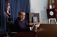 a man sitting at a desk with a microphone in front of him and several flags behind him