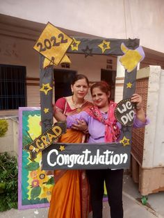 two women standing next to each other in front of a building holding a sign that says congratulations