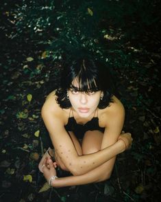 a woman is sitting in the woods with her hands on her chest and looking at the camera