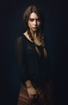 a woman with long hair standing in front of a dark background wearing a black shirt and striped skirt