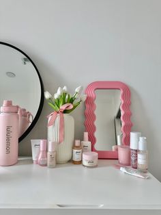 a white dresser topped with lots of beauty products and a pink vase filled with flowers