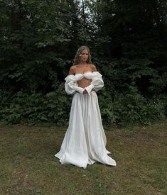 a woman wearing a white dress standing in the grass with trees behind her and looking off to the side