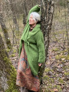 an older woman leaning against a tree in the woods wearing a green sweater and scarf