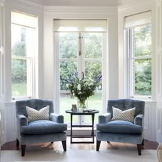 two chairs and a table in front of three windows with blue couches, one is facing the other