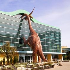 a giant dinosaur statue in front of a large building with a green roof and glass windows