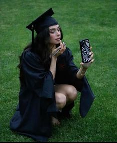 a woman in a graduation cap and gown kneeling on the grass while holding a cell phone