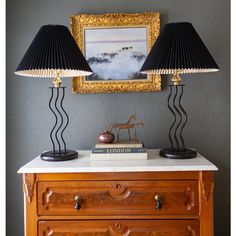 two lamps sitting on top of a wooden dresser next to a painting and lamp shade