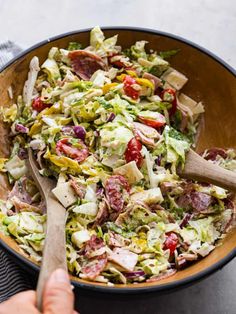 a bowl full of salad with wooden spoons in it and someone's hand on the side