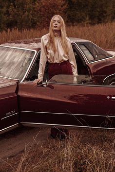 a woman leaning on the hood of a red car in a field with tall grass