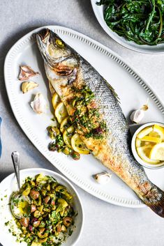 a fish and some vegetables on a white plate with silver spoons next to it