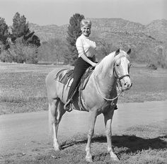 a woman riding on the back of a white horse down a dirt road next to a field