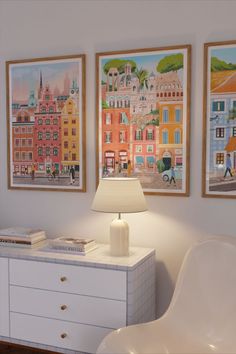 three paintings hang on the wall next to a white dresser with a lamp and chair