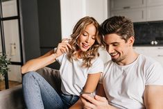 a man and woman sitting on a couch looking at a cell phone while they laugh