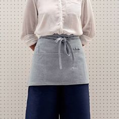 a woman standing in front of a white wall wearing an apron with pockets on it