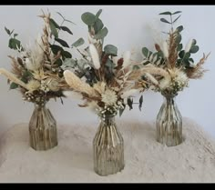 three vases with dried flowers and greenery in them on a white blanketed surface