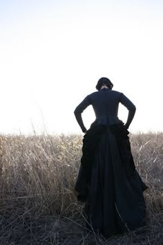 a woman in a long black dress standing in tall grass with her back to the camera