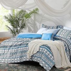 a bed covered in blue and white sheets next to a potted plant on top of a rug