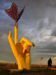 a large yellow sculpture with a purple butterfly on it's head, in front of a cloudy sky